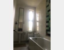 Helles Badezimmer mit weiß gefliesten Wänden, einer Badewanne rechts im Vordergrund, einem Waschbecken mit Spiegel links und einem Fenster mit hellem Tageslicht im Hintergrund. Rechts an der Wand befindet sich ein Abschnitt mit grünen und schwarzen Mosaikfliesen.