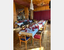 Ein festlich eingedeckter Raum mit einer langen Tafel, weißen Tischdecken, roten Stoffservietten und mehreren gedeckten Stühlen in verschiedenen Farben. Im Hintergrund sind ein Buffet, eine rote Vorhangwand sowie eine dekorative Decke mit Netzen und einem Kronleuchter zu sehen.