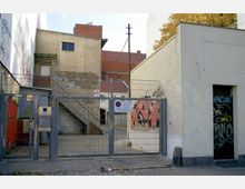 Metalltor mit Zaun in einer urbanen Umgebung, dahinter ein betonierter Innenhof mit gelber Backsteinwand und Außentreppe. Die Wände sind teils mit Graffiti versehen, und auf dem Tor sind Schilder mit Park- und Zutrittsbeschränkungen angebracht.