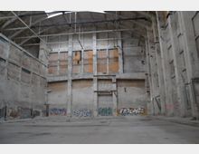 Weitläufiger, leerer Innenraum einer verlassenen Industriehalle mit hohen Betonwänden, teilweise offengelegten Ziegeln und großen Fenstern, die teilweise mit Holz verkleidet sind. Die unteren Bereiche der Wände sind mit Graffiti verziert, und an der Decke verlaufen Metallträger.