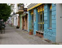 Straßenszene in einem urbanen Wohn- und Geschäftsviertel mit bunt bemalten Gebäuden, darunter ein blaues Haus mit Schaufenstern und gelben Markisen. Links ist ein schmaler Gehweg mit Bäumen, Fahrrädern und kleinen Geschäften zu sehen.