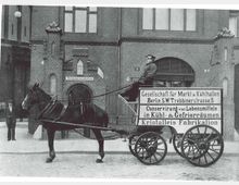 Historische Aufnahme eines Backsteingebäudes mit einem Pferdewagen im Vordergrund, auf dem "Gesellschaft für Markl & Kühlhallen, Berlin S.W. Trebbinerstraße 5" sowie Hinweise zur Lebensmittelkühlung und Eisherstellung stehen. Ein Mann sitzt auf dem Wagen, während ein weiterer Mann neben dem Torbogen des Gebäudes steht.