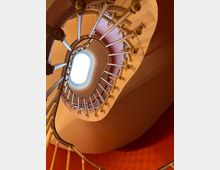 Blick von unten auf ein gewendelt gebautes Treppenhaus mit ovaler Öffnung in der Decke, die Tageslicht durch ein Fenster hereinlässt. Die Wände sind in Rot- und Beigetönen gehalten, und das Geländer besteht aus hellen Metallstreben.