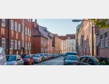 Straßenszene in einer Stadt mit Kopfsteinpflaster, gesäumt von mehrstöckigen Backsteingebäuden und Wohnhäusern in verschiedenen Farben. Autos parken auf beiden Straßenseiten, und die Szene wird von einer Straßenlaterne sowie vereinzeltem Laub im Vordergrund ergänzt.