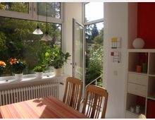 Esszimmer mit großen Fenstern, durch die ein Garten mit Bäumen sichtbar ist. Auf der Fensterbank stehen Zimmerpflanzen in weißen Töpfen, daneben ein Holztisch mit gestreifter Tischdecke und Holzstühlen; im Hintergrund ein Regal mit Dekorationen und Wandbildern in geometrischen Mustern.