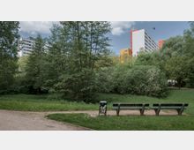 Städtischer Park mit einer Wiese, Bäumen und Büschen im Vordergrund, daneben ein Weg mit Sitzbänken und einem Mülleimer. Im Hintergrund sind mehrstöckige Wohngebäude mit farbigen Fassaden zu sehen.