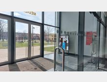 Eingangsbereich eines modernen Gebäudes mit Glaswänden und -türen, durch die ein Außenbereich mit Gehweg, Bänken und Bäumen sichtbar ist. Im Inneren steht rechts eine Pinnwand mit verschiedenen Aushängen, und ein Heizkörper ist an der Wand montiert.