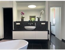 Modernes Badezimmer mit schwarzem Fliesenboden und schwarzer Mosaikrückwand, davor ein schwebender weißer Waschtischunterschrank mit ovalem Aufsatzwaschbecken und großer Spiegelwand. Im Vordergrund ist der Rand einer freistehenden Badewanne zu sehen, rechts führt eine Tür in einen hellen Nebenraum mit Fenster.