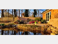 Ein gepflegter Garten mit einem kleinen Teich im Vordergrund, umgeben von Natursteinen und Pflanzen. Im Hintergrund steht eine Ziegelmauer mit einer eingelassenen Nische, daneben ein Haus aus roten Ziegelsteinen mit großen Fenstern.