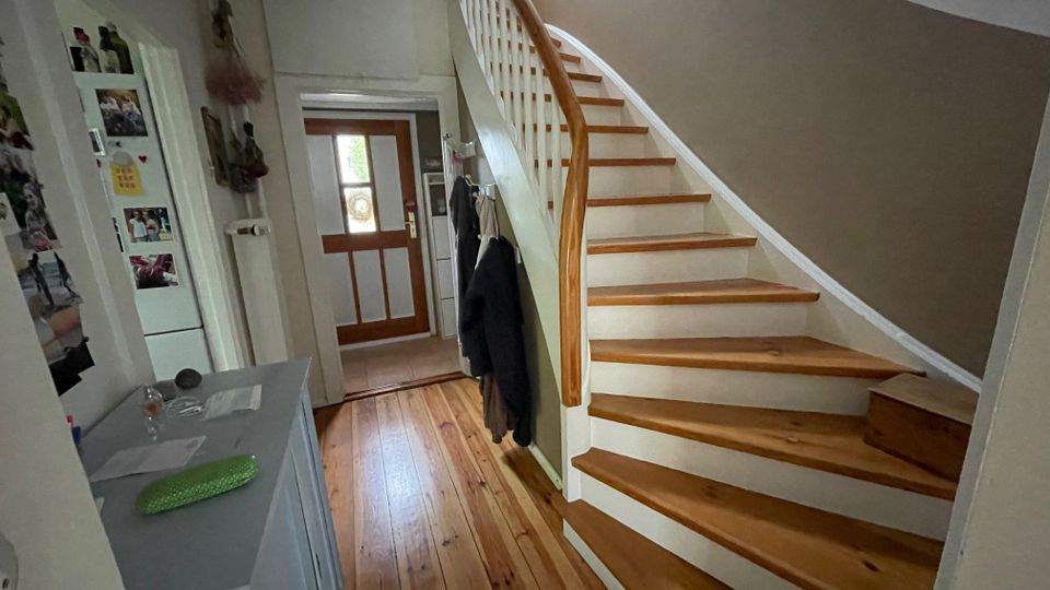 Flur im Eingangsbereich eines Hauses mit Holzfußboden und einer geschwungenen Holztreppe mit weißem Geländer, die nach oben führt. Am Ende des Flurs steht eine Haustür mit Glaseinsatz; seitlich sind eine Kommode und Wandhaken für Jacken zu sehen.