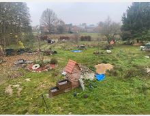 Grüne Wiese mit unebener Fläche und verstreuten Baustellenmaterialien, wie Sandhaufen, Ziegelsteine und Planen; im Vordergrund ein gemauerter Brunnen mit Ziegeldach. Im Hintergrund sind Bäume, ländliche Gebäude und ein kiesiger Weg sichtbar.