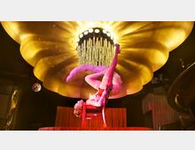 Eine Theaterbühne mit einer Performer*in in einem roten Kostüm, die auf einem Stuhl kopfüber balanciert. Im Hintergrund ein großer dekorativer, goldener Kronleuchter in Blütenform und schummriges Bühnenlicht.