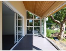 Moderner Wintergarten mit großen Glasfronten, hellem Holzdeckendach und grauen Bodenfliesen, angrenzend an einen Garten mit Bäumen und Wiese.