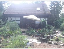 Ein kleines Fachwerkhaus mit rotem Ziegeldach, umgeben von einem dicht bepflanzten, ländlichen Garten. Im Vordergrund sind ein Steinweg, verschiedene Pflanzen, Gartenwerkzeuge und ein Sonnenschirm zu sehen.