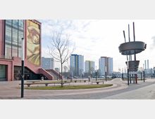 Moderne Stadtansicht mit einem Platz im Vordergrund, der von Bänken und einem kahlen Baum geziert wird. Links befindet sich ein Gebäude mit einer großen Wandgestaltung, rechts ein Kunstwerk aus Metall, im Hintergrund mehrere farbige Hochhäuser und ein Parkplatz.