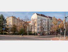 Straßenszene mit mehrstöckigen Wohngebäuden im klassischen Stil, teils mit Balkonen, flankiert von Bäumen mit Herbstlaub. Im Vordergrund eine Kreuzung mit Verkehrsampeln und Radwegmarkierungen.