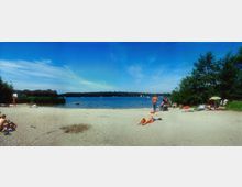 Strandabschnitt an einem See mit sandigem Ufer, im Hintergrund dichter Wald und einige Segelboote auf dem Wasser. Menschen entspannen auf Handtüchern und Liegen, einige schwimmen oder nutzen Schlauchboote, während am Rand Bäume und Büsche das Ufer säumen.
