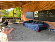 Eine überdachte Terrasse mit einer Wand aus rotem Backstein, auf der eine große Tafel mit der Aufschrift „Hallo Team, let's rock 2022!“ angebracht ist. Der Boden besteht aus festem Sand, es gibt einen großen Sofabereich mit Decken und Kissen sowie bunte Stühle und Tische; im Hintergrund sind grüne Büsche und Bäume sichtbar.