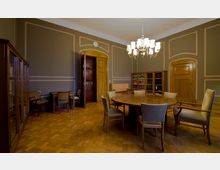 Tagungsraum mit Holzmöbeln, darunter ein runder Tisch und mehrere gepolsterte Stühle. Der Raum hat Parkettboden, hohe Wände mit dekorativen Zierleisten und Schränke mit Büchern, sowie eine große Deckenleuchte.