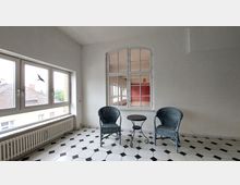 Heller Raum mit großem Fenster, durch das Dächer und Bäume draußen sichtbar sind. In der Ecke stehen zwei blaue Flechtstühle mit einem kleinen runden Tisch davor, der Boden ist mit schwarz-weißen Fliesen gemustert, und eine innenliegende Sprossenwand zeigt ein weiteres Zimmer mit roter Wand.