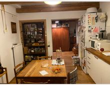 Gemütliche Küche in einem rustikalen Stil mit freiliegenden Holzbalken an der Decke. Zu sehen sind ein Holzesstisch mit Stühlen, ein Regal mit Vorräten, ein Kühlschrank mit Magneten sowie eine Mikrowelle auf einer Küchenzeile; im Hintergrund ist ein Schrank zu erkennen.