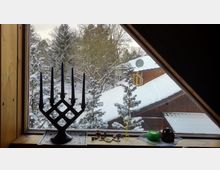 Blick aus einem Dachfenster auf eine schneebedeckte Landschaft mit Bäumen und einem Holzhaus mit Satellitenschüssel. Auf der Fensterbank steht ein schwarzer Kerzenständer mit blauen Kerzen, daneben liegen kleine Dekorationsobjekte und ein grünes Teelichtglas.
