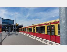 Bahnhof mit moderner Glasfassade und roter S-Bahn auf dem Gleis. Der Bahnsteig ist mit Sitzbänken, Abfallbehältern und Beschilderungen ausgestattet, daneben ein blauer Himmel im Hintergrund.