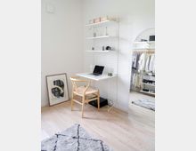 Helle, minimalistisch eingerichtete Ecke eines Wohnraums mit einem kleinen weißen Schreibtisch, Holzstuhl und Regal. Ein Spiegel reflektiert ein offenes Ankleidezimmer mit hängender Kleidung, während ein Teppich und ein gerahmtes Bild den Raum ergänzen.