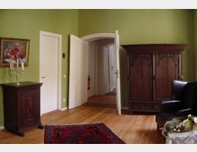 Zimmer mit grünen Wänden, Holzboden und klassischen Möbeln, darunter ein großer dunkler Holzschrank, ein kleines Sideboard mit Kerzenständer und ein Ledersessel. Eine geöffnete weiße Tür führt in einen angrenzenden Raum, und im Vordergrund liegt ein gemusterter Teppich.