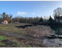 Eine ländliche Uferlandschaft mit zwei kleinen Fachwerkhäuschen im Hintergrund, einer grasbewachsenen Fläche und mehreren ungenutzten Holzplattformen. Rechts im Bild fließt ein kleiner Fluss oder Kanal, neben dem ein leeres Boot auf einem schmalen, betonierten Zugang liegt.