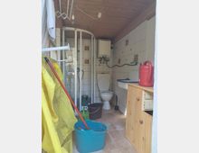 Schmaler, gefliester Sanitärraum mit Holzdecke, Duschkabine links und Toilette am Ende; an der rechten Wand stehen ein kleines Waschbecken mit Unterschrank, ein Heizkörper und ein Wandgerät. Im Vordergrund sind Putzutensilien wie Eimer, Mop und Gießkanne sowie eine rote Thermoskanne auf dem Schrank zu sehen.