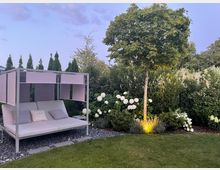 Moderner Gartenbereich mit Rasenfläche, einem weißen Daybed mit Rahmen und Sonnensegel auf Kiesbett sowie dichtem Grün aus Hecken und Sträuchern. Rechts steht ein kugelförmig geschnittener Baum mit Bodenbeleuchtung, umgeben von weißen Hortensienblüten.