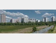 Weites Feld mit hohem Gras und vereinzelten Büschen im Vordergrund, ein Zaun verläuft diagonal. Im Hintergrund stehen mehrere weiße und graue Wohnhochhäuser, umgeben von einer Baumreihe. Der Himmel ist teilweise bewölkt.