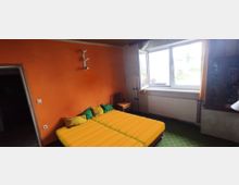 Ein einfaches Schlafzimmer mit orange gestrichener Wand und gelb-grüner Einrichtung. Zwei Einzelbetten mit gelben Decken stehen nebeneinander, ein großes Fenster mit Vorhängen lässt Tageslicht herein, und an der Wand hängt eine kleine Lampe.