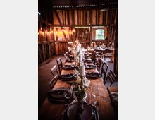 Eine festlich gedeckte Tafel in einer rustikalen Scheune mit Holzwänden und -balken, geschmückt mit Kerzen, Pflanzenarrangements und Geschirr. Lichterketten hängen an den Wänden, und ein kleines Fenster mit Gitter verleiht dem Raum Tageslicht.