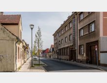 Straßenszene mit Wohnhäusern aus Putz und Backsteinfassaden, auf der linken Seite ein altes Gebäude mit Ziegeldach und Graffiti. Ein Gehweg mit jungen Bäumen und Straßenlaternen verläuft parallel zu einer asphaltierten Straße.