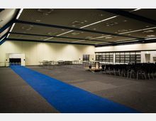 Großer Veranstaltungsraum mit grauem Teppichboden, einer blauen Läuferbahn in der Mitte und weiß-beigen Wänden. Der Raum ist mit Stühlen auf der rechten Seite, leeren Tischen und einer kleinen Bühne ausgestattet; die Decke weist ein Rastermuster mit eingebauten Leuchten auf.