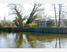 Moderne Gebäude mit großen Glasfronten am Ufer eines ruhigen Flusses, umgeben von hohen, teils bewachsenen Bäumen und einer mit Efeu bedeckten hölzernen Uferbefestigung. Im Wasser schwimmen zwei Enten.