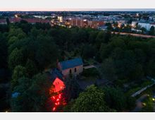 Luftaufnahme eines Parks bei Dämmerung mit dichtem Baumbestand, einer Kirche mit blauem Dach und angrenzendem rot beleuchteten Gebäude. Im Hintergrund sind Wohn- und Bürogebäude sowie beleuchtete Straßen der Stadt sichtbar.