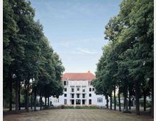 Weiße Villa mit rotem Ziegeldach, symmetrischer Fassade und Balkonen, umgeben von hohen Bäumen, die einen Weg durch einen grasbedeckten Garten einrahmen. Der Himmel ist klar und blau.