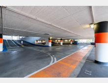 Ein moderner, überdachter Parkbereich mit markierten Parkplätzen und Fahrspuren, beleuchtet durch Deckenleuchten. Sichtbar sind eine Rampe zur oberen Ebene, farblich markierte Säulen in Weiß, Orange und Schwarz sowie vereinzelte parkende Fahrzeuge.