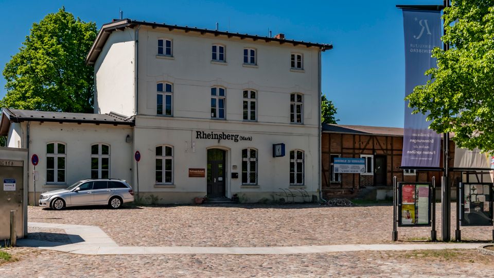 Vorderansicht eines kleinen Bahnhofsgebäudes mit weißer Fassade und der Aufschrift „Rheinsberg (Mark)“, davor ein gepflasterter Vorplatz. Rechts stehen Informationsvitrinen und ein Fahnenmast unter Bäumen, seitlich schließt ein niedriger Holzbau an.