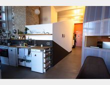 Kleine, moderne Küche in einem offenen Wohnraum mit sichtbarer Ziegelsteinwand, weißen Geräten, Holzarbeitsplatten und einer Wandregalablage. Im Hintergrund führt ein Flur mit gelblicher Beleuchtung zu einer rosa Tür, rechts ist ein Arbeitsbereich mit transparenten Trennwänden sichtbar.