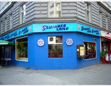 Eckgebäude mit blauer Fassade und dem Schriftzug „Slumberland“ auf einem blauen Leuchtkasten über den Fenstern, daneben ein Aufkleber mit der Marke „Schneider Weisse“. Die Fenster geben den Blick auf ein Innenrauminterieur mit Pflanzen und Möbeln frei.