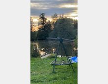 Eine grüne Wiese mit einer Metall-Gartenschaukel im Vordergrund, dahinter ein kleiner See, umgeben von Bäumen, mit einem Steg am Ufer und einem Sonnenuntergang am Himmel.