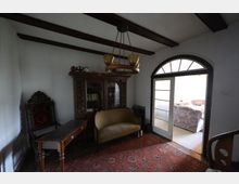 Ein kleines Wohnzimmer mit Holzbalkendecke, antikem Möbelstil und Teppichboden. Zu sehen sind ein verziertes Bücherregal, ein gelbes Sofa, ein Holztisch und ein gepolsterter Stuhl. Eine doppelflügelige Glastür mit Rundbogen führt in einen angrenzenden Raum.