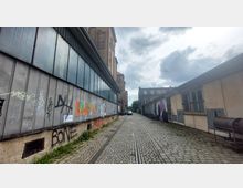 Schmale gepflasterte Gasse mit Schienen in der Mitte, flankiert von industriell wirkenden Gebäuden. Links ein mehrstöckiges Gebäude mit Metallfassade, versehen mit Graffiti, rechts ein niedrigeres Gebäude mit vergitterten Fenstern und Wandmalereien.
