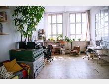 Heller Raum mit großen Fenstern, der als Arbeits- und Wohnbereich genutzt wird. Der Raum ist mit Pflanzen, einem Schreibtisch, Regalen, einem braunen Ledersessel und verschiedenen Büromöbeln eingerichtet.