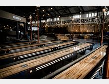Großer Innenraum einer modernen Bierhalle mit Industriecharakter, geprägt von Backsteinwänden, hohen Decken mit freiliegenden Stahlträgern und Lichtgirlanden. Im Vordergrund sind mehrere Shuffleboard-Tische und im Hintergrund Sitzbereiche mit Holzmöbeln zu sehen, sowie eine Wand mit der Aufschrift „Geboren in Schottland. In Deutschland gebraut.“