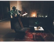 Gemütliche Lounge mit dunklem Sofa, Polstersessel und weißem Beistelltisch auf einem roten Teppich mit orientalischem Muster. Dekorative Zimmerpflanzen, eine Stehlampe und Kerzen auf einer schwarz lackierten Ablage erzeugen eine warme Beleuchtung in einem Raum mit Betonwänden.